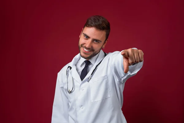 白い医療用ガウンを着た若いハンサムな現代の医者は、赤い背景のスタジオに立っています。医学部の学生研修生. — ストック写真