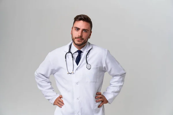 Jovem médico moderno bonito em um vestido médico branco fica no estúdio em um fundo branco. Estudante estagiário de uma universidade de medicina . — Fotografia de Stock