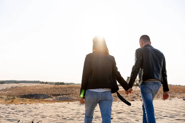 Joven pareja europea vestida chaqueta de cuero y pantalones vaqueros azules caminando desierto sosteniendo las manos vista desde la parte posterior . — Foto de Stock