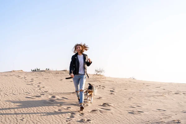 Een jong blank meisje ergens in een leren jas en blauwe jeans loopt langs het zandstrand met haar beagle dog — Stockfoto