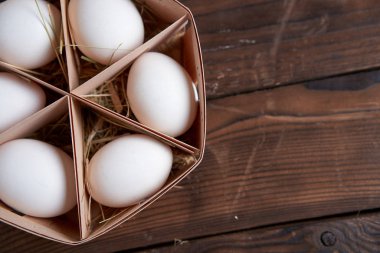 White chicken eggs lie in round wooden basket which stands on a dark wooden table.