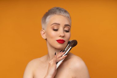 Portre makyözünü kapat. Kursları hazırla. Kişisel görünüm ana sınıflandırması kavramı. Moda uzmanı. Kadın elinde makyaj fırçası tutuyor. Turuncu arkaplanda stüdyo çekimi