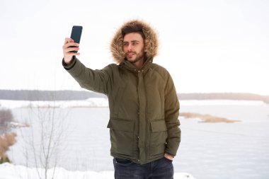 Yakışıklı, sakallı, beyaz, açık havada duran, kürk kapüşonlu, donmuş göl mutlu, çekici Avrupalı adam akıllı telefon fotoğrafçısı Kış Bülteni macerasında selfie çekiyor.