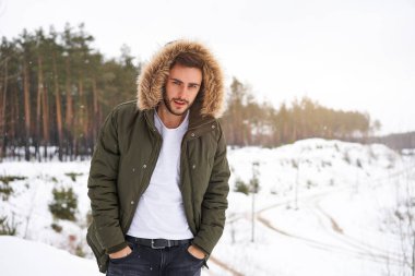 Yakışıklı, sakallı, beyaz bir adam kış mevsimi ormanında dışarıda kürk kapüşonlu duruyor. Çekici stil sahibi Avrupalı adam, karlı Noel ormanlarında yürüyor.                       