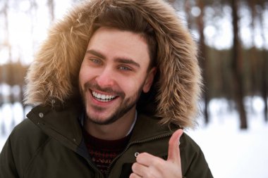 Yakışıklı, sakallı, beyaz, açık havada duran tüylü, kış mevsimi ormanı. Çekici stil sahibi Avrupalı adam karlı Noel ormanlarında yürüyor Noel tatilleri boş zamanlarında başparmak işareti yapıyor.