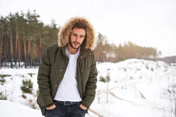 Yakışıklı, sakallı, beyaz bir adam kış mevsimi ormanında dışarıda kürk kapüşonlu duruyor. Çekici stil sahibi Avrupalı adam, karlı Noel ormanlarında yürüyor.                       