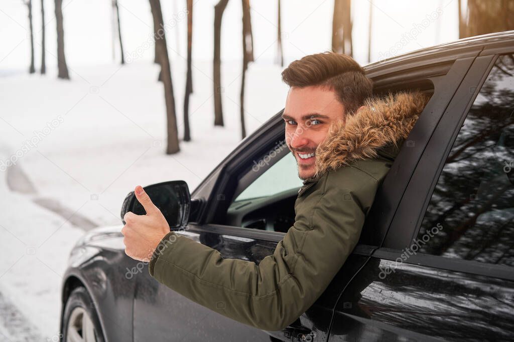 Young adult attractive Caucasian man sits at the wheel of his car sunny winter day shoeing thumbs up gesture. Wintertime road trip. Happy smiling hipster guy sitting in car and looking window. 