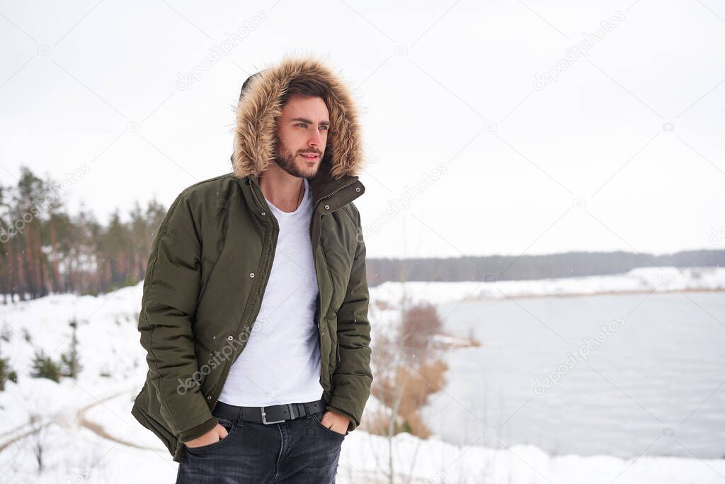 Handsome bearded young caucasian man standing outdoors fur hood in winter season forest. Attractive stylish european guy walking snowy christmas woodland Season holiday leisure                       