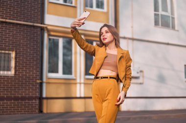 20 yaşında, beyaz saçlı, şık turuncu takım elbiseli güzel beyaz bir kız sokakta dururken telefonda selfie çekiyor. Moda blogcusu kendi fotoğraflarını çekiyor. 