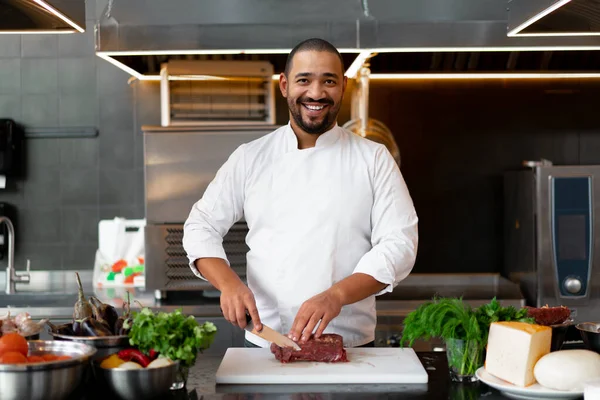 Yakışıklı genç Afrikalı şef restoranın profesyonel mutfağında duruyor ve et ve peynir sebzelerinden oluşan bir yemek hazırlıyor. Üniformalı bir adamın portresi metal bir bıçakla et keser..
