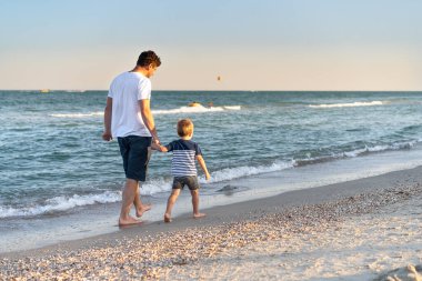 Küçük oğlu olan genç beyaz bir baba deniz kıyısında sıcak bir yaz günü yürüyor. Yaz tatili konsepti. Baba oğul arkadaşlığı. Ailenle sahilde mutlu bir çocukluk. Metin için boş alan