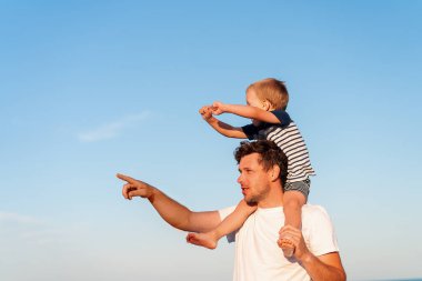 Young Caucasian dad with little son walk warm summer day along the sea coast. Summer family vacation concept. Friendship father and son. Father holding son on his shoulders at the beach
