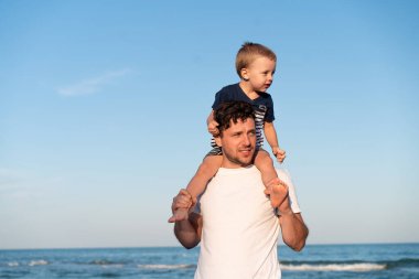 Young Caucasian dad with little son walk warm summer day along the sea coast. Summer family vacation concept. Friendship father and son. Father holding son on his shoulders at the beach
