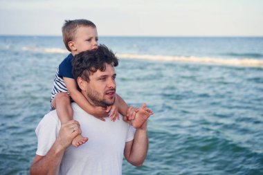 Young Caucasian dad with little son walk warm summer day along the sea coast. Summer family vacation concept. Friendship father and son. Father holding son on his shoulders at the beach