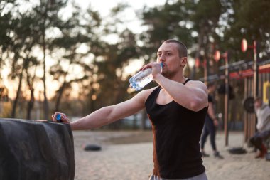 Sporcu, tekerlek lastiğine dayanmış plastik şişe egzersizi içer. Genç, yakışıklı, beyaz bir adam, doğaya çok yüklendikten sonra dinleniyor, bir lastiğin yanında duruyor, soğuk su içiyor..
