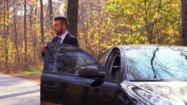 Un beau jeune homme caucasien portant un costume d'affaires debout près de voiture noire sur la nature . — Video