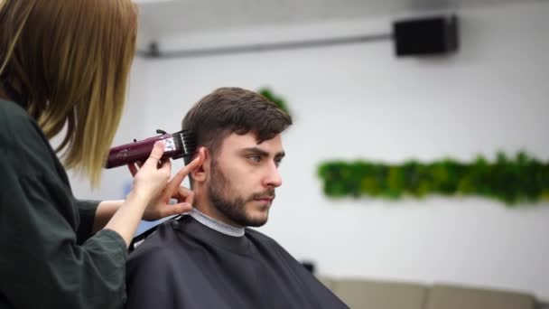 Hombre guapo de ojos azules sentado en la peluquería. Peluquera Peluquera cortándole el pelo. Peluquero femenino. — Vídeo de stock