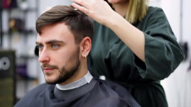 Przystojny niebieskooki facet siedzi w salonie fryzjerskim. Fryzjerka obcinająca mu włosy. Kobieta fryzjer. — Wideo stockowe