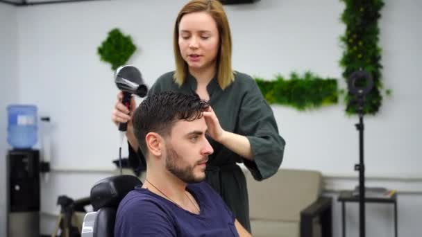 Bonito homem de olhos azuis sentado na barbearia. Cabeleireiro Cabeleireiro Mulher cortando seu cabelo. Barbeiro fêmea. — Vídeo de Stock