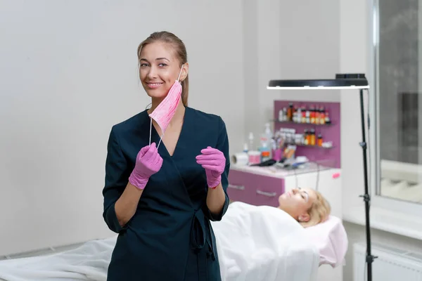 Cosmetology cabinet client lies on couch Beautician stands pink protective medical mask glove and smiles Preparation for the procedure permanent eyebrow makeup Full coronavirus protection covid-19