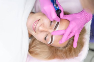 Kozmetik dolabı müşterisi kanepede yatıyor. Güzellik uzmanı kaşlara kalıcı mürekkep uygular. Kalıcı kaş makyajı prosedürü. Kapatın. Güzellik endüstrisi Pembe eldivenler