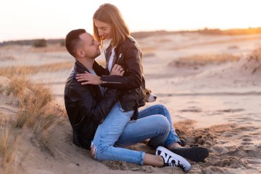 Siyah deri ceket ve kot pantolon giymiş güzel beyaz bir çift. Bir adam kumların üzerine oturur ve ona sarılır. Sevgili genç çift aşıkları güzel günbatımı arka planını öper..