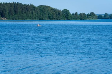 Büyük, geniş bir nehir ve ortasında iki balıkçı ile bir tekne; diğer tarafında ağaçlar, evler ve orman vardır