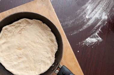 Kızartma tavası ve hamur bir kesim tahtasının üzerinde duruyor. Masaya un serpiştirilmiş.