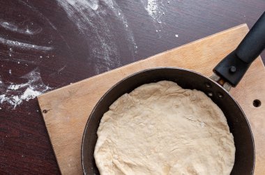 Kızartma tavası ve hamur bir kesim tahtasının üzerinde duruyor. Masaya un serpiştirilmiş.