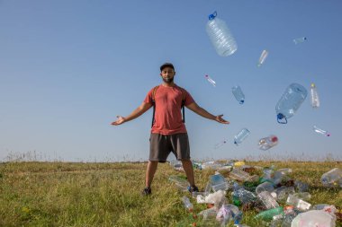 Kırmızı tişörtlü genç bir adam yeşil bir tepede duruyor. Plastik çöp ve şişelerle dolu.