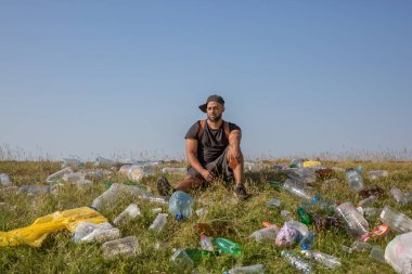 Beyzbol şapkalı genç bir adam bir dizini yeşil bir yamaçta plastik şişelerin ve çöplerin arasında büker..