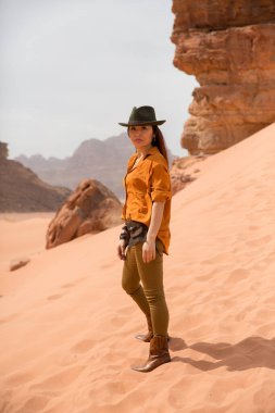 Kovboy çizmeleri ve şapkası olan güzel Asyalı kız kumlu bir yamaçta duruyor. Arka planda çöl ve dağlar. Mart 2020 'de Ürdün' de çekildi.