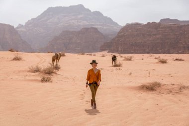 Şapkalı ve çizmeli bir kız çölde, develerin arkasında, kumların üzerinde yürüyor. Wadi Rum çölünde vuruldu.