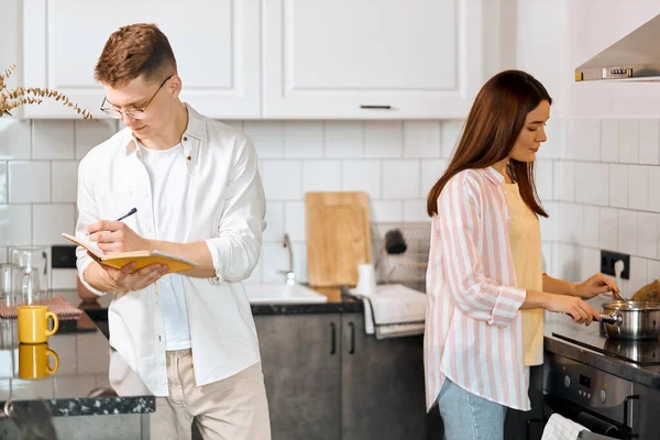 Jovem mulher cozinhar mingau de cereal enquanto seu marido fazendo um projeto . — Fotografia de Stock