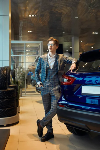 Junger eleganter Mann mit gekreuzten Beinen, der auf seinem blauen Auto steht — Stockfoto