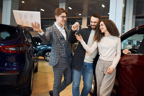 Successful professional cheeful dealer is handing auto keys to a young couple — Stockfoto