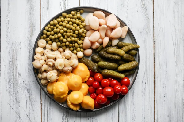 Fermented food in a plate on a white textural wooden table. Cucumbers, garlic, mashrooms, tomatoes, green peas, patissons, champignons. National Russian food pickles in brine close up and copy space