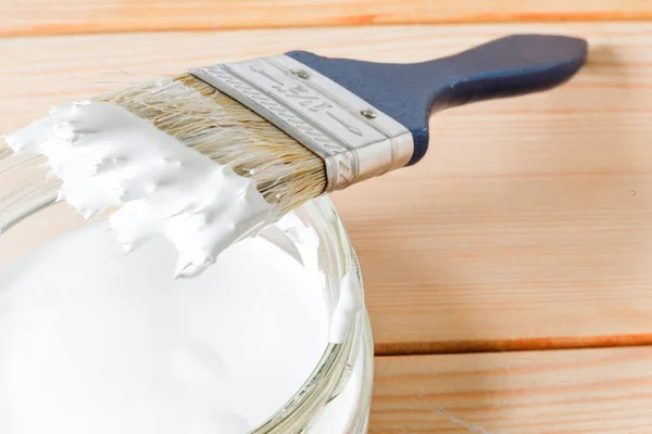 Blauwe kwast in witte verf ligt op een glazen pot met verf, tegen de achtergrond van lichte planken. Close-up, selectieve focus — Stockfoto