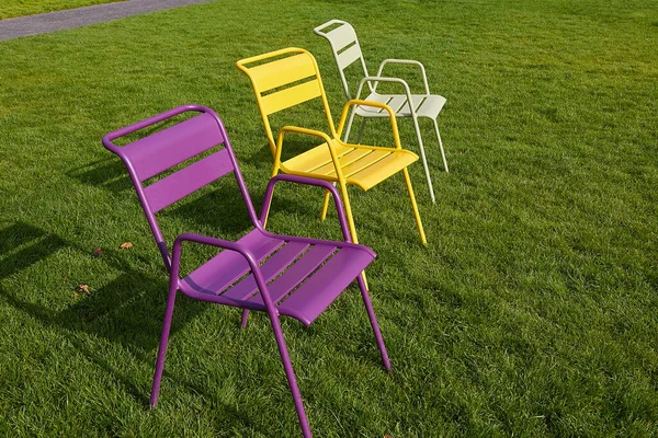 Three multi-colored metal chairs stand on a green lawn in the rays of the setting sun. Image of the urban environment. — Stock Photo, Image