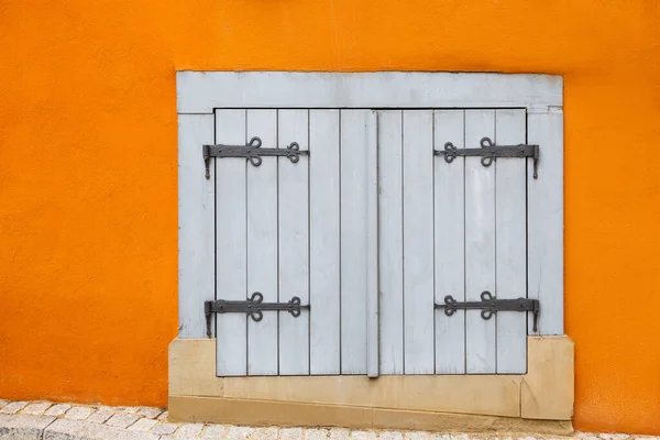 Una piccola finestra bianca è chiusa da persiane in legno con cerniere decorative su una parete arancione brillante . — Foto Stock