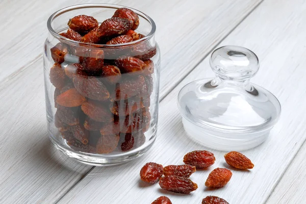 Dried rosehip berries in a glass jar and several scattered berries on a light wooden table. The concept of traditional medicine, treatment with natural medicinal plants. Close-up. — Stock Photo, Image