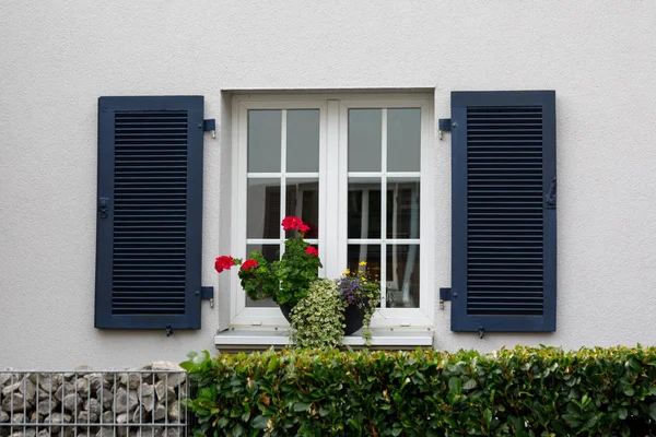 Ett fönster med vita bågar, mörkblå fönsterluckor och blommor på fönsterbrädan. En fasad av ett traditionellt europeiskt hus på landet. — Stockfoto