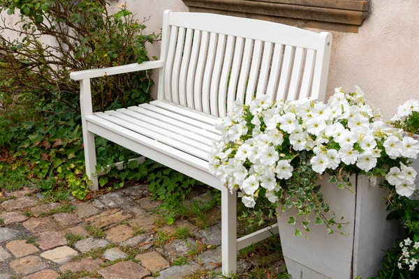 Sui ciottoli vicino ai fiori di petunia bianca che crescono in una grande scatola di legno si trova una comoda panca di legno bianca. Il comfort di una piccola città — Foto Stock