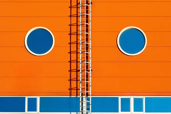 A fragment of a orange modern facade of an industrial building, warehouse or exhibition center with two symmetrical round windows and a metal staircase. Graphic Association with a persons face — Stock Photo, Image