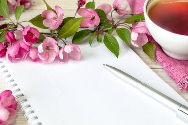 Kahvaltı için bir fincan çay, boş bir defter ve beyaz masada pembe elma çiçekleri. Masada çalışan bir kadın. Üst görünüm düz yatış biçimi.