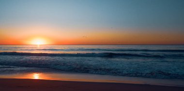 Soyut deniz manzarası. Okyanusun üzerinde gün batımı. Mavi okyanus dalgaları, kumsal, renkli gökyüzü ve ufukta batan güneş.