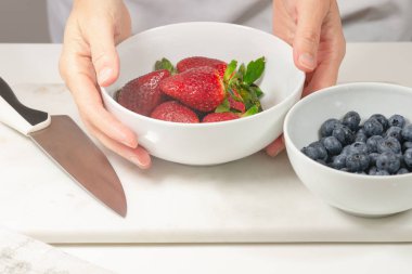 Strawberries and blueberries. Close up of fresh organic berries in a bowl, woman hands, white background. Woman preparing dessert