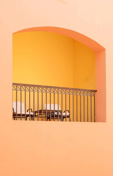 Minimalist picture of yellow wall of building and terrace with chairs and table for rest in the center.