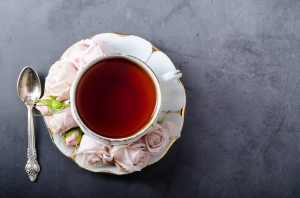Çay saati fon. Üstten alta beyaz porselen çay fincanı, yumuşak pembe güller ve koyu gri arka planda güzel bir çay kaşığı.. — Stok fotoğraf