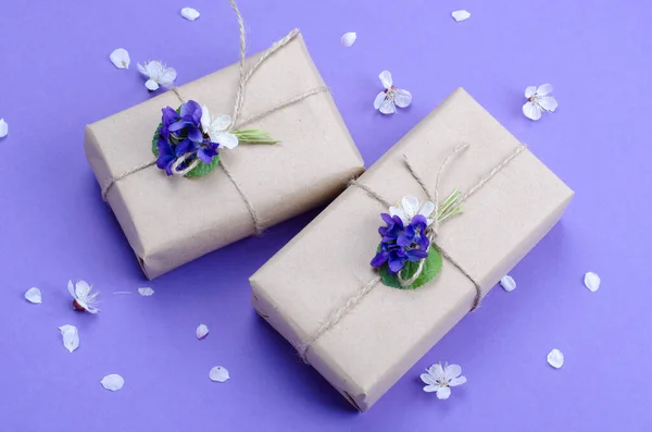 Mooie geschenkdozen verpakt in eenvoudige bruine ambachtelijke papier versierd met levende bloemen van violet op een violette achtergrond. — Stockfoto